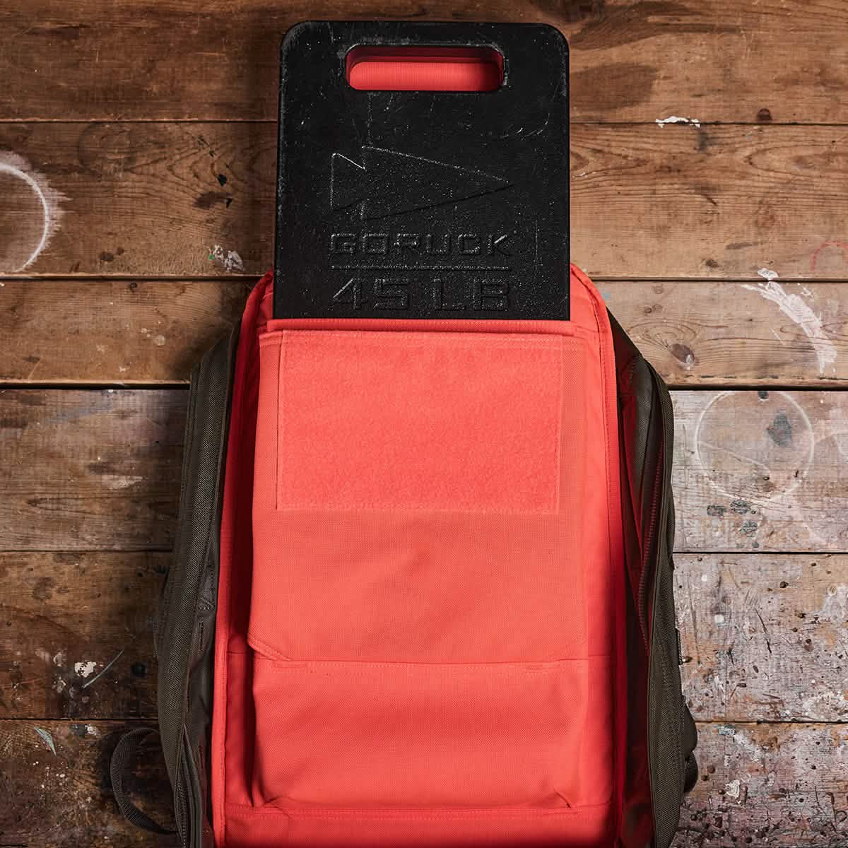 GORUCK 45 LB weight plate in orange backpack on rustic wooden surface, men's gear
