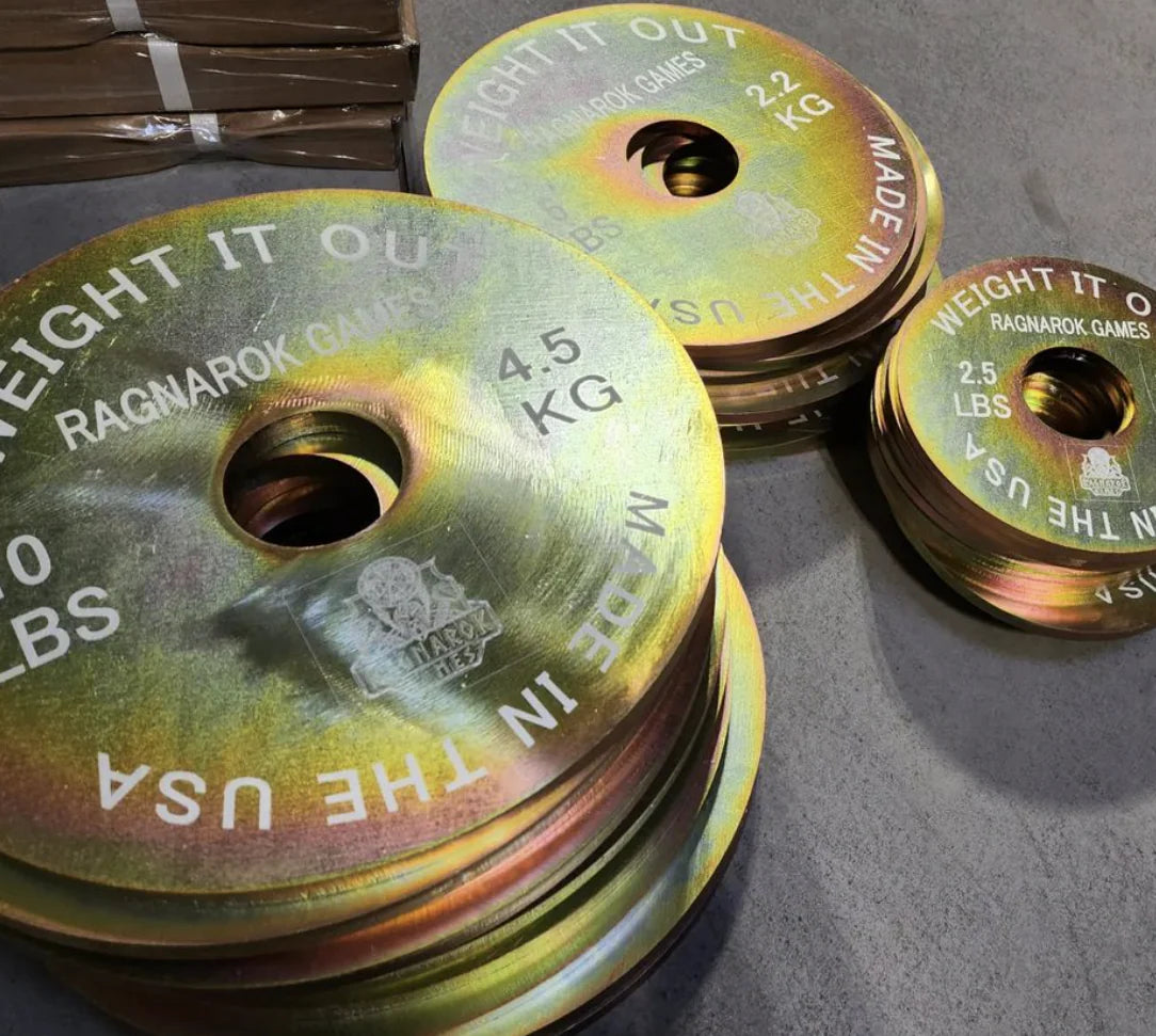 Stack of custom metal weight plates labeled 'Weight It Out' and 'Ragnarok Games' on gym floor