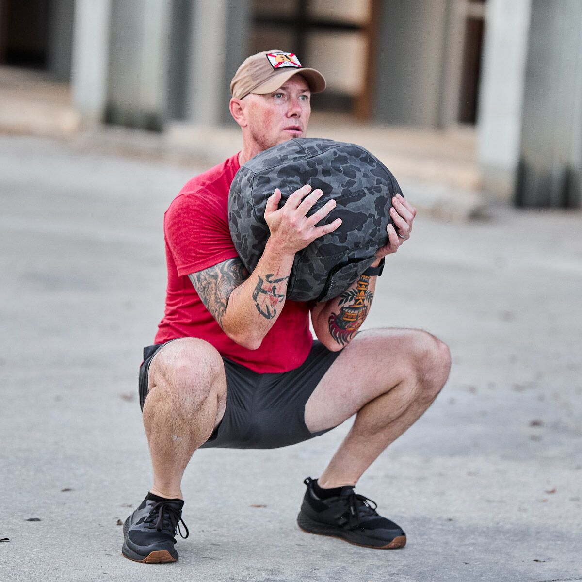 Man in red shirt and shorts doing outdoor squat exercise with a camo fitness sandbag