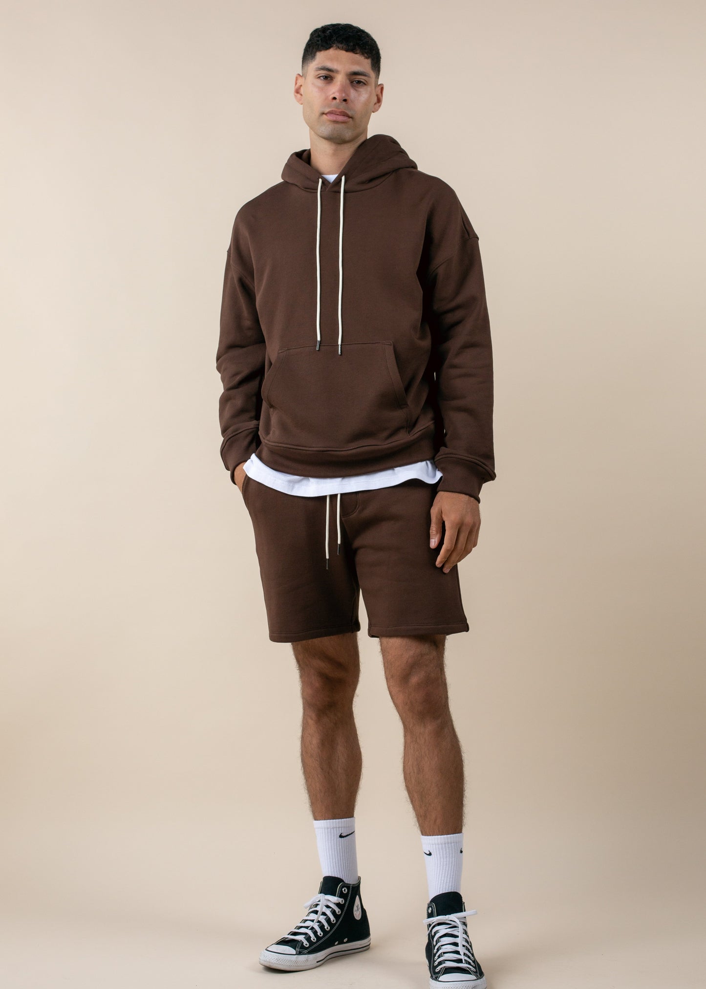 Man in brown hoodie and shorts, white socks, and black sneakers, posing against beige background for MANN men's fashion marketplace.