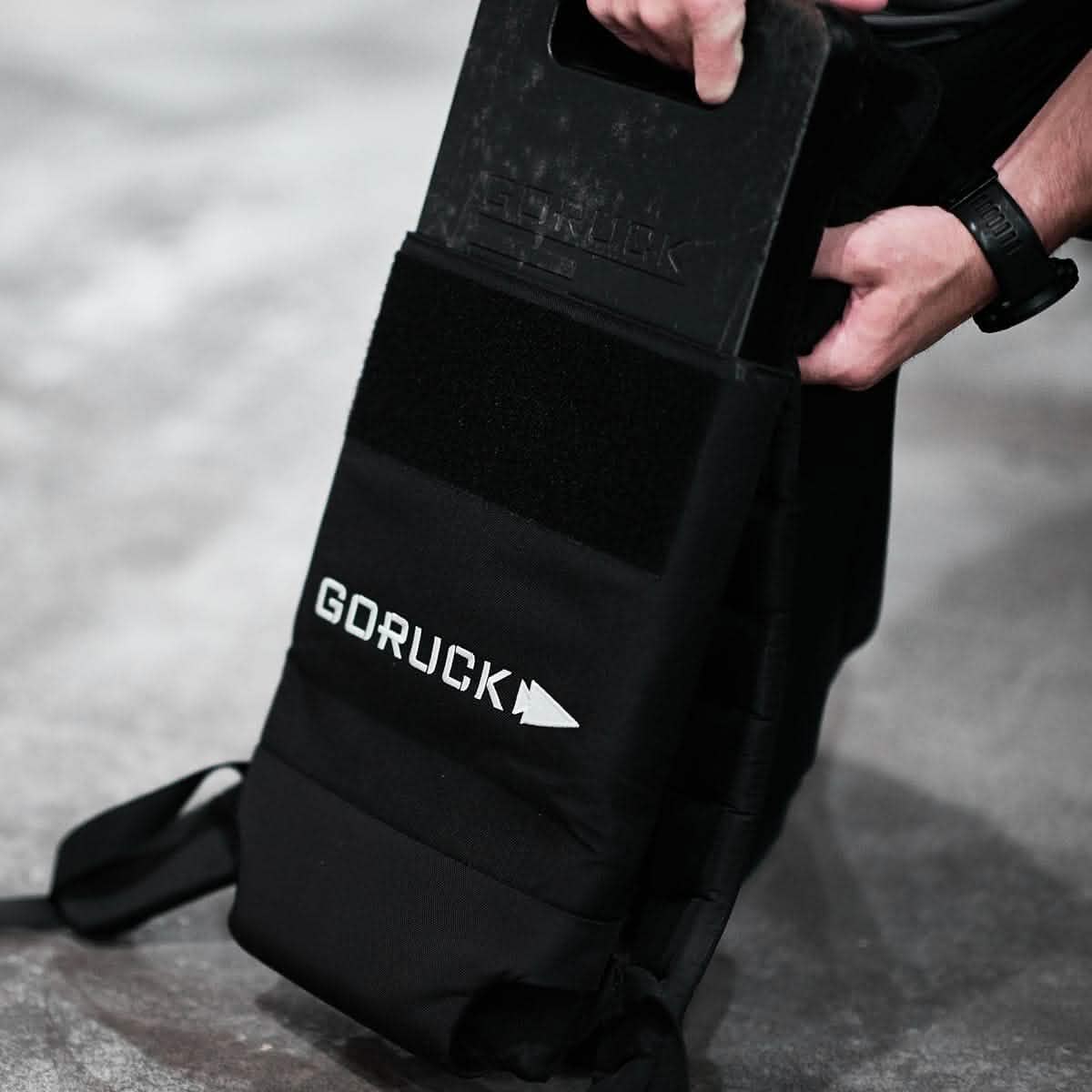 Man placing GORUCK 45LB weight plate in black fitness backpack on concrete floor