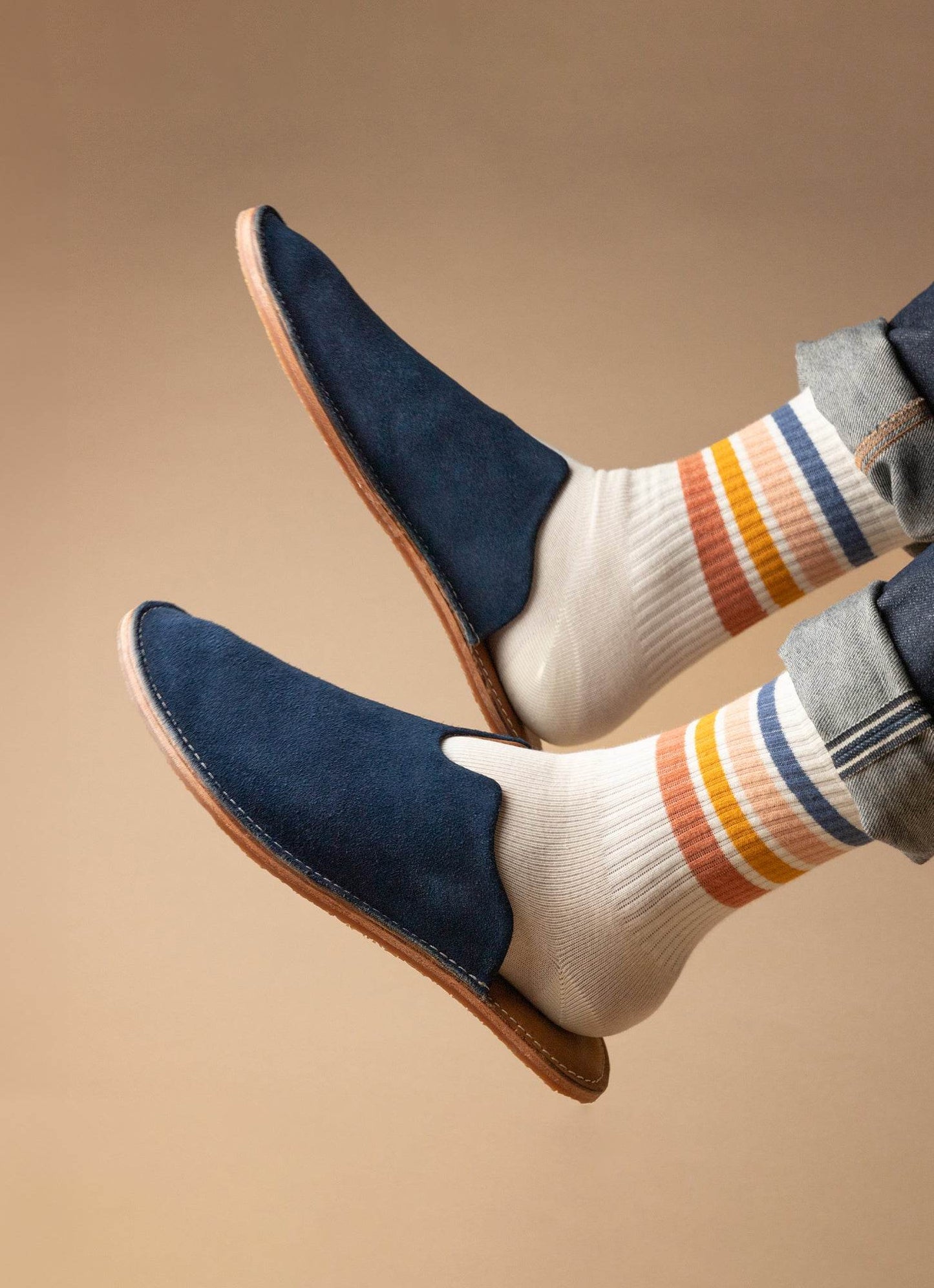 Men's navy suede slip-on shoes with striped socks, rolled denim, on neutral background