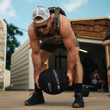 Fit man in camouflage vest lifting 60lb GORUCK medicine ball outside gym, men's fitness gear