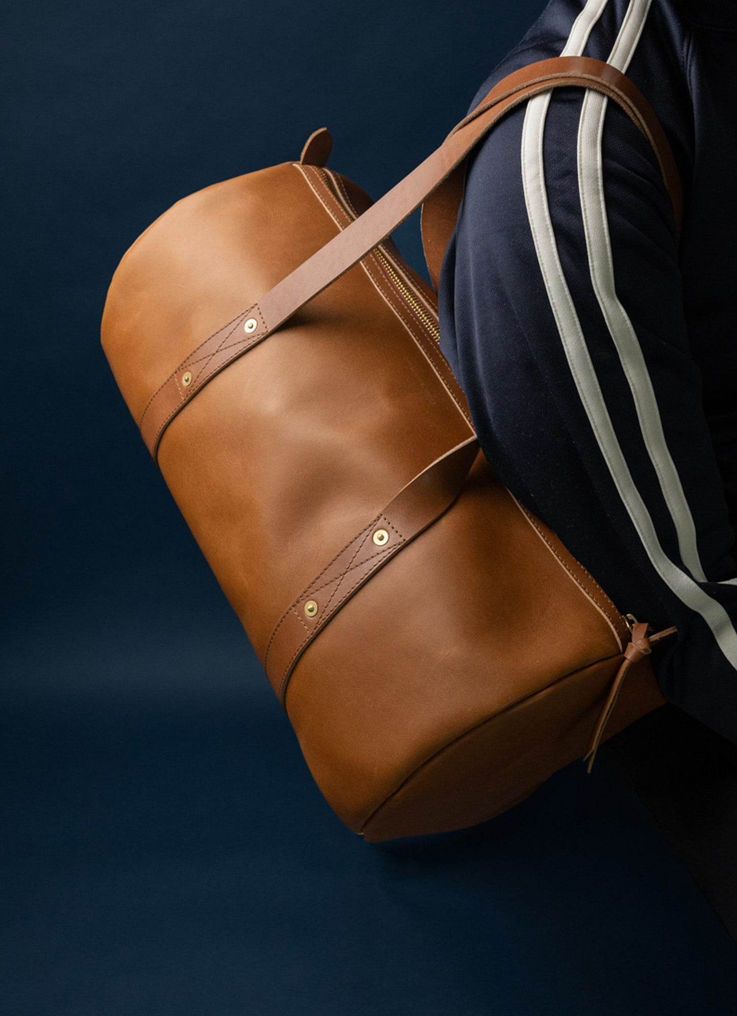 Men's brown leather duffel bag with shoulder strap, worn over navy athletic jacket