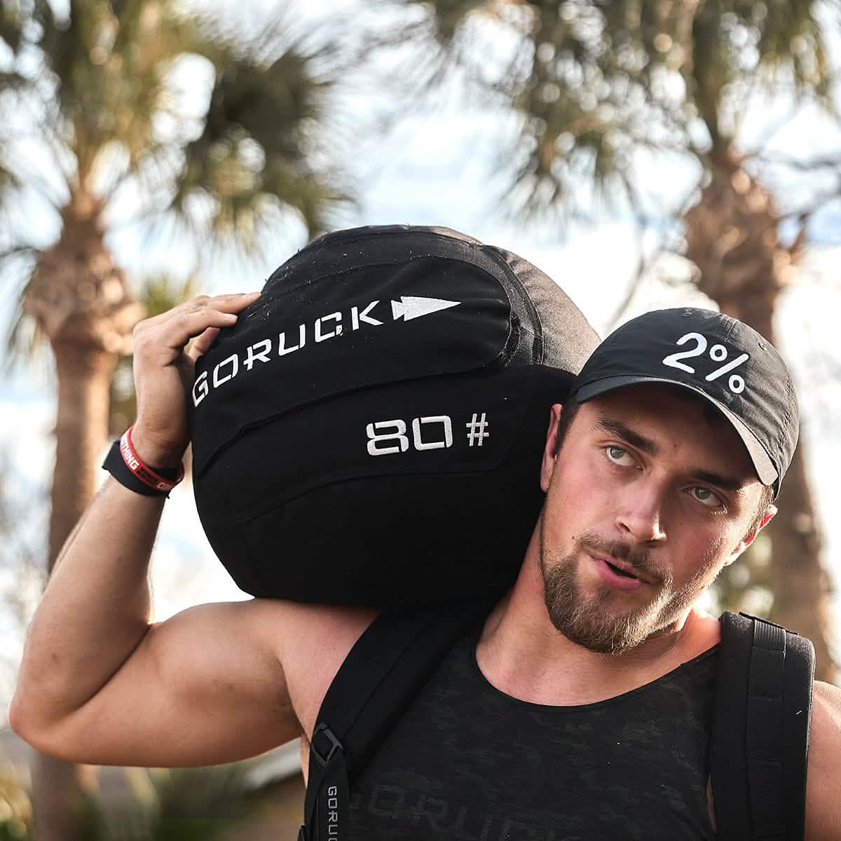 Man carrying GORUCK 80lb sandbag outdoors, wearing black tank top and 2% cap, palm trees behind