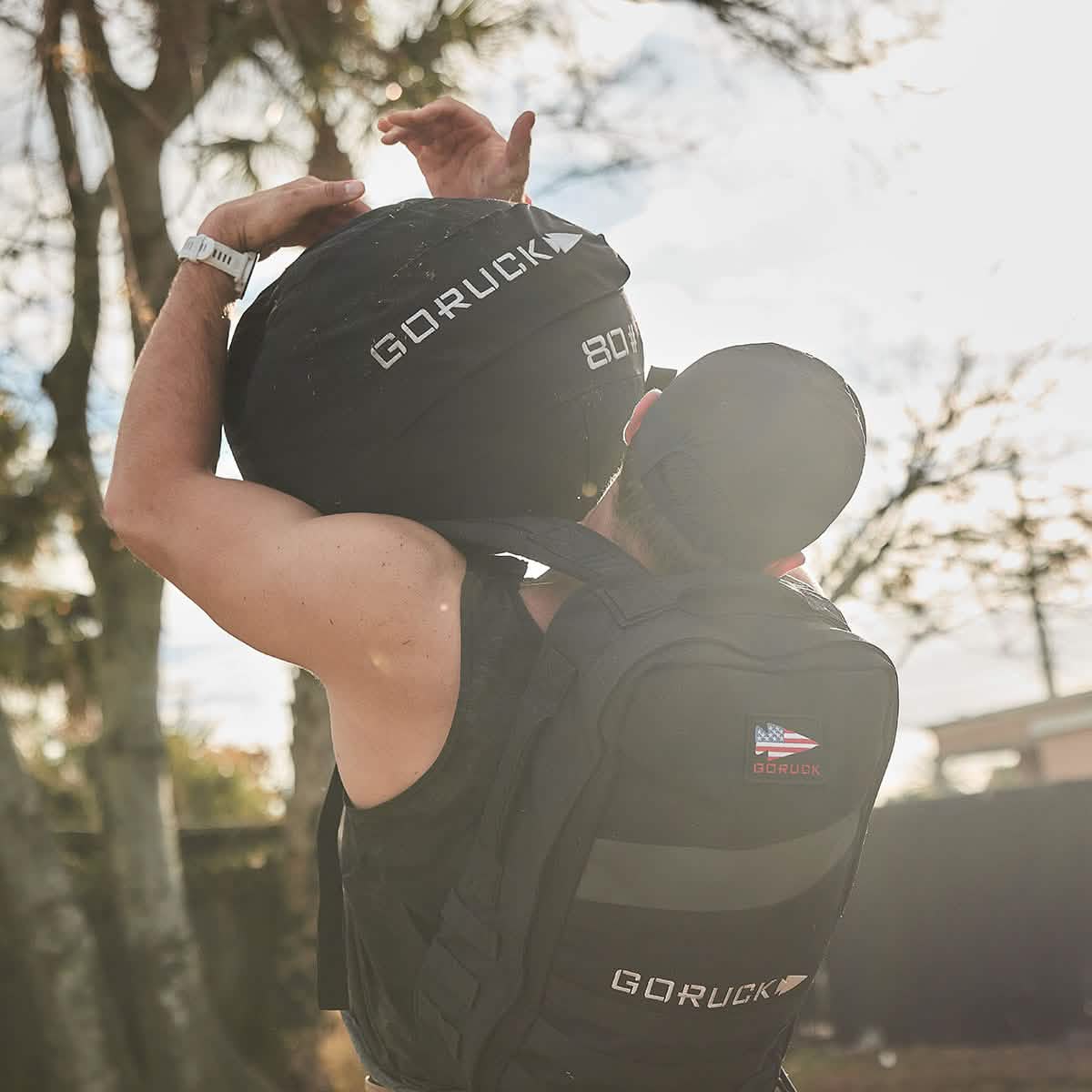 Man outdoors lifting GORUCK 80lb sandbag, wearing GORUCK backpack and black cap