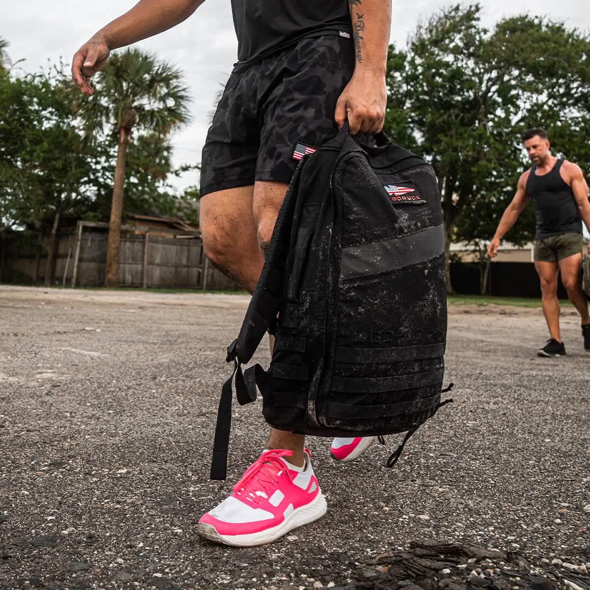 Man in workout shorts and bright sneakers holding a black tactical backpack outdoors