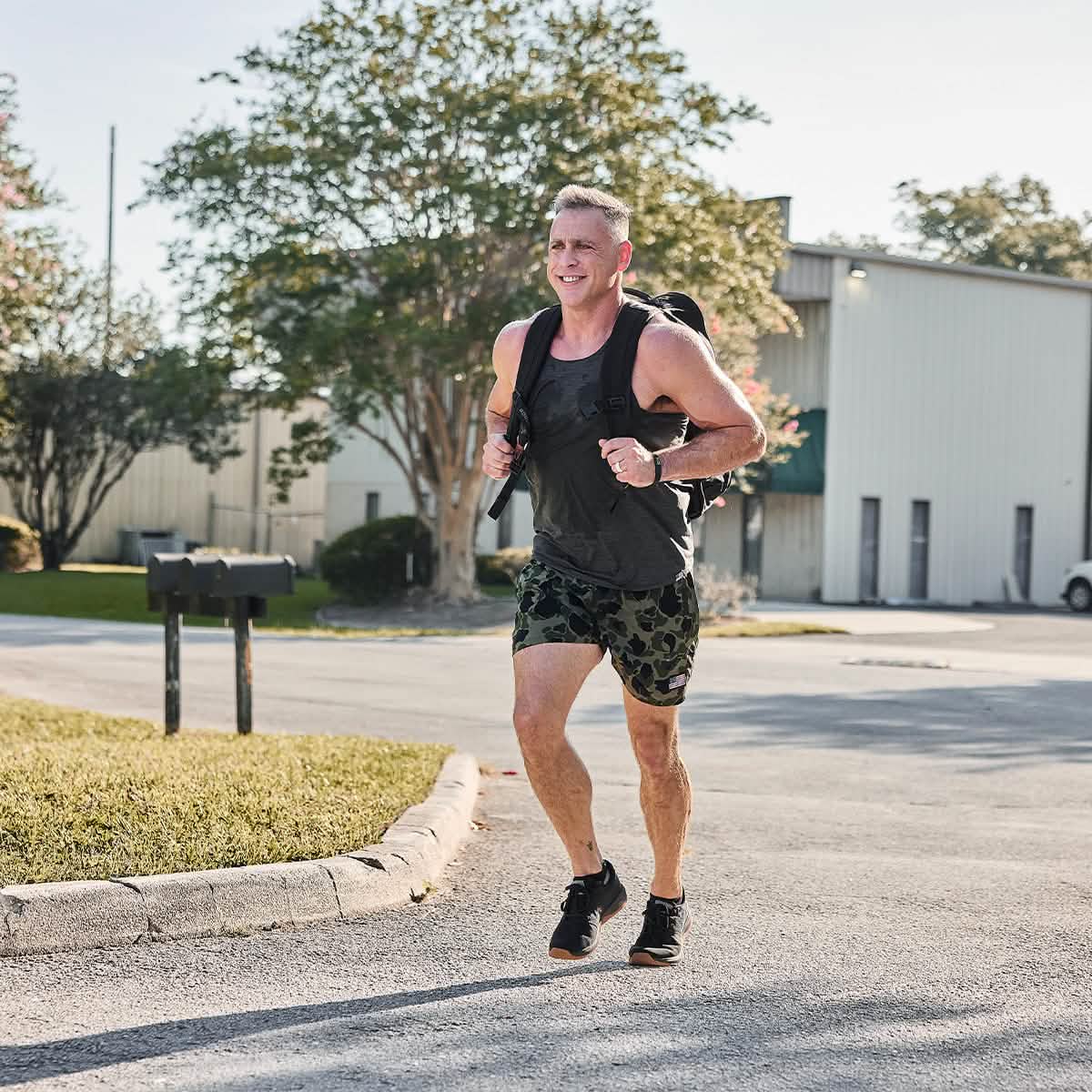 Athletic man running outdoors in black tank top, camo shorts, and backpack, showcasing men's active lifestyle fashion.