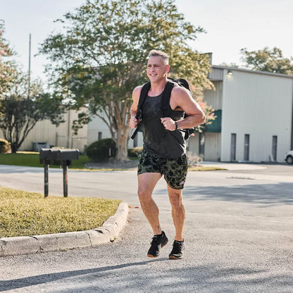 Athletic man running outdoors in black tank top, camo shorts, and backpack, showcasing men's active lifestyle fashion.