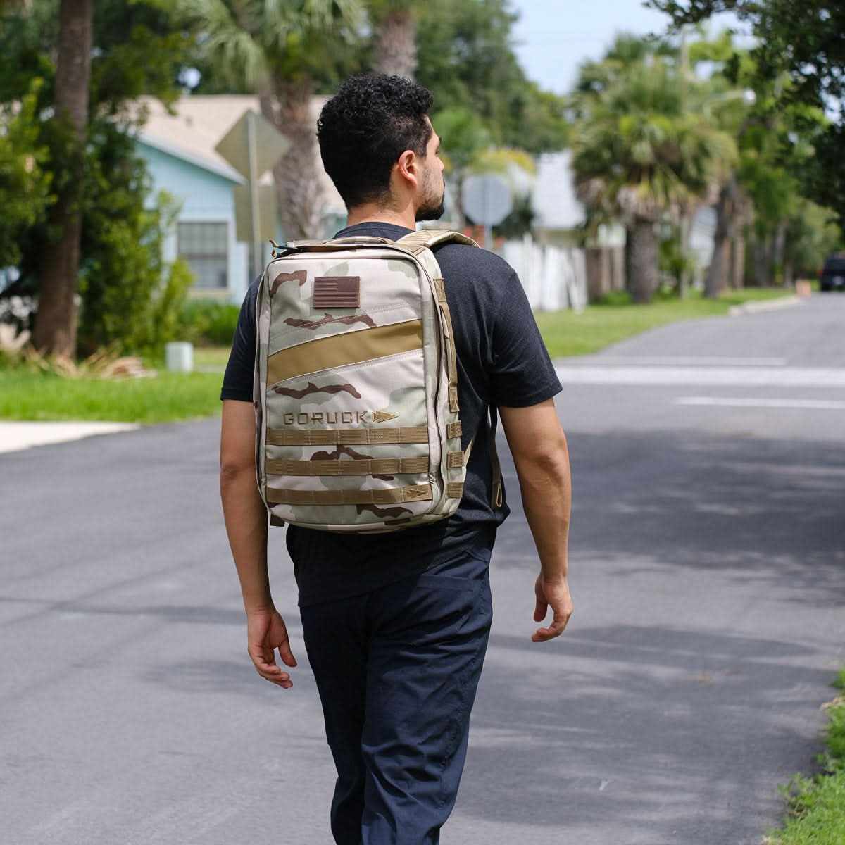 Man wearing a camo GORUCK backpack walking outdoors on a sunny neighborhood street.