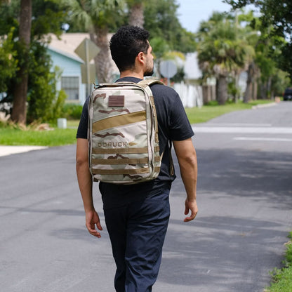 Man wearing a camo GORUCK backpack walking outdoors on a sunny neighborhood street.