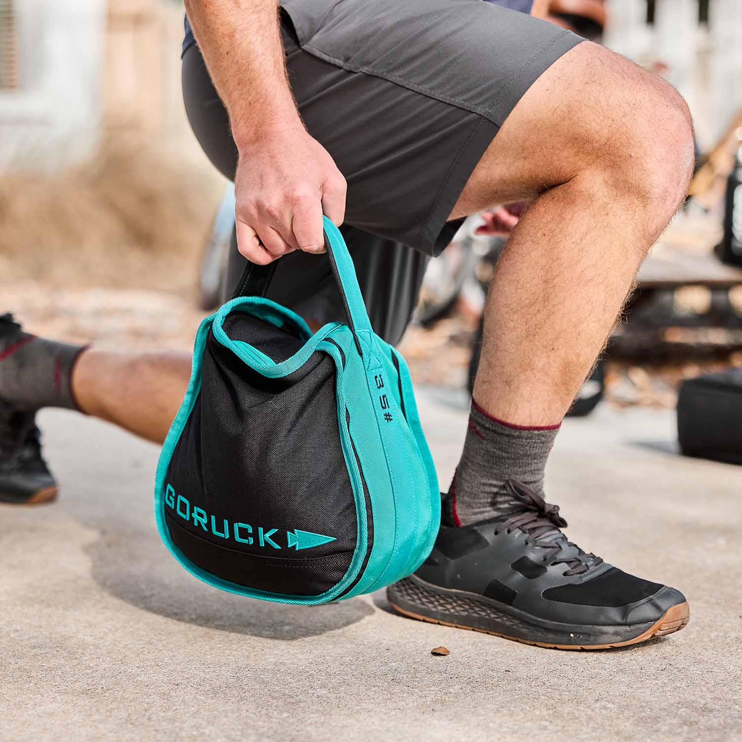 Man exercising outdoors holding a black and teal GORUCK kettlebell, wearing athletic gear