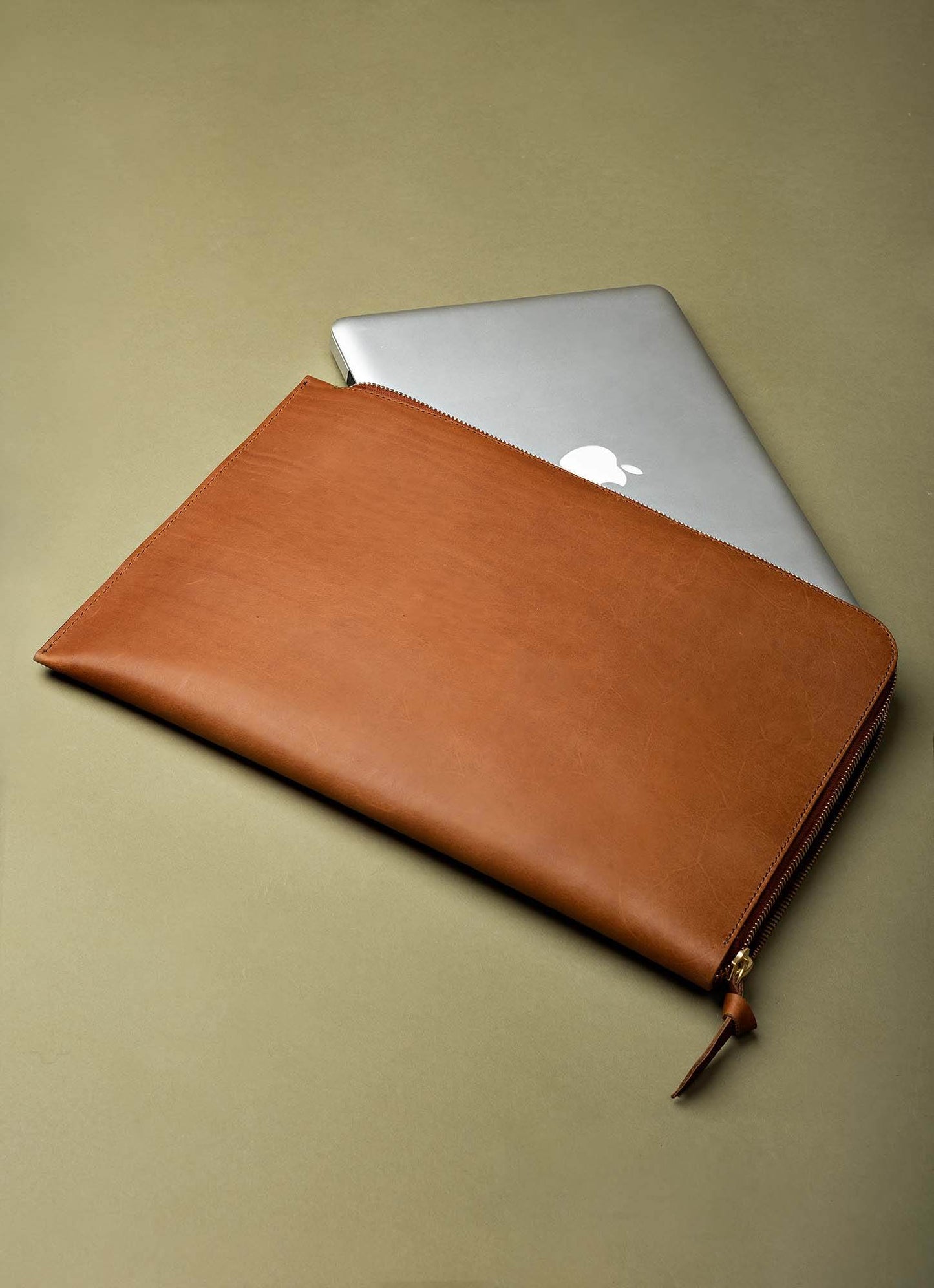 Slim brown leather laptop sleeve with zipper, partially covering silver laptop on neutral background