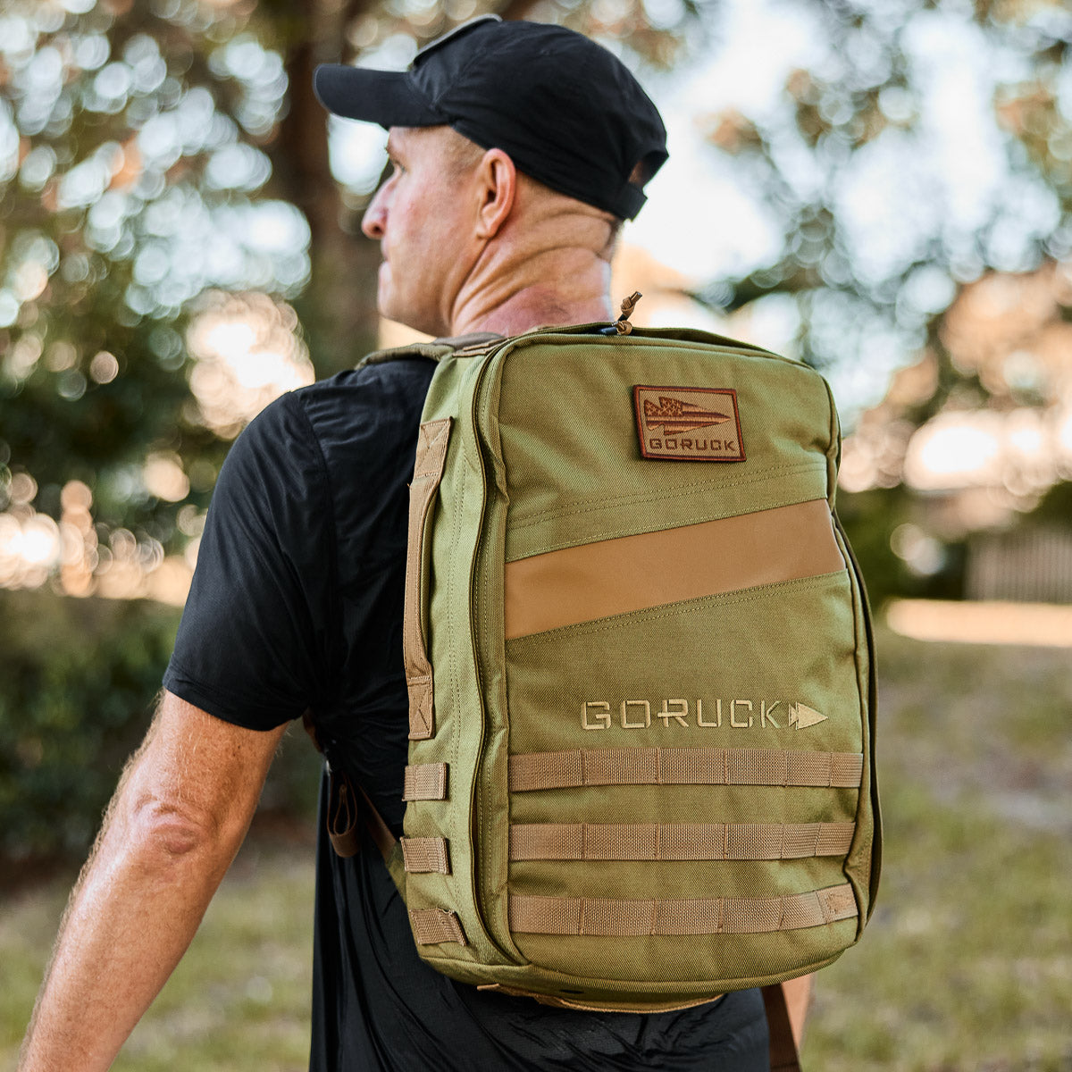 Man outdoors wearing olive GORUCK backpack, black shirt and cap, curated men's lifestyle gear