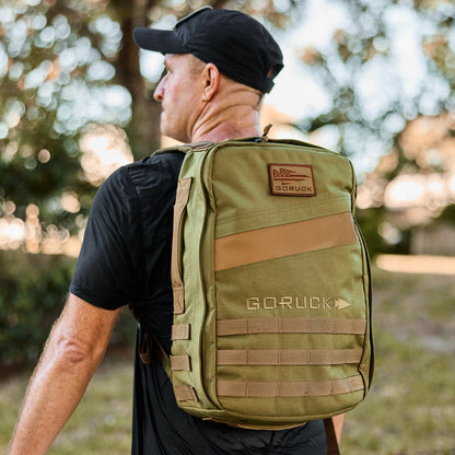 Man outdoors wearing olive GORUCK backpack, black shirt and cap, curated men's lifestyle gear