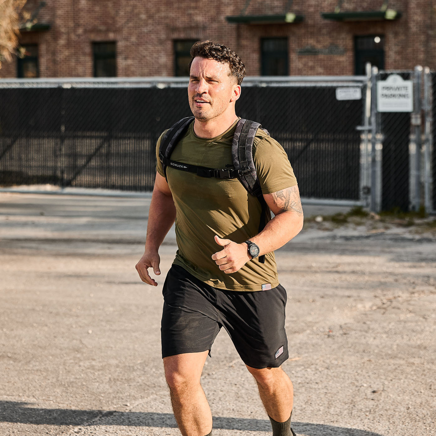 Athletic man jogging outdoors in black shorts and olive t-shirt with backpack, urban background