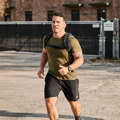 Athletic man jogging outdoors in black shorts and olive t-shirt with backpack, urban background