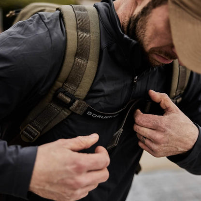 Man adjusting olive tactical backpack strap, wearing a cap and outdoor jacket