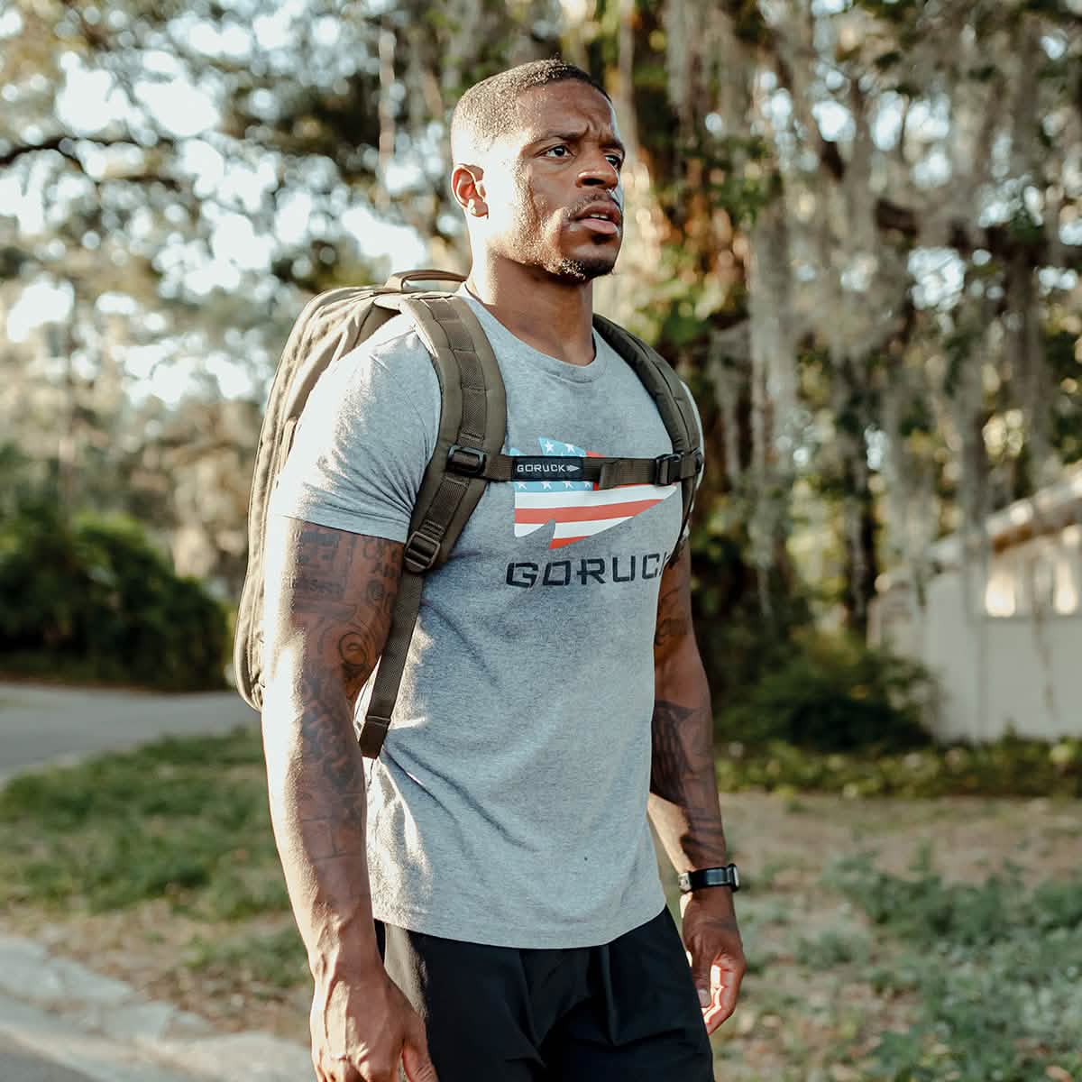 Man outdoors wearing a gray GORUCK t-shirt and olive green backpack, fitness lifestyle