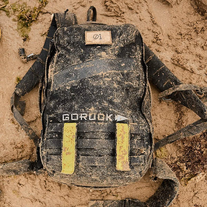 GORUCK tactical backpack with yellow straps, covered in sand, on an outdoor dirt surface
