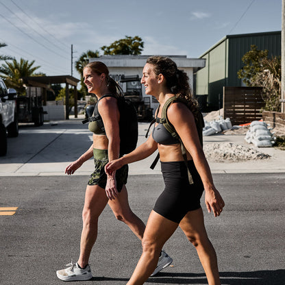 Two women in athletic wear and backpacks walking outdoors in a sunny urban setting