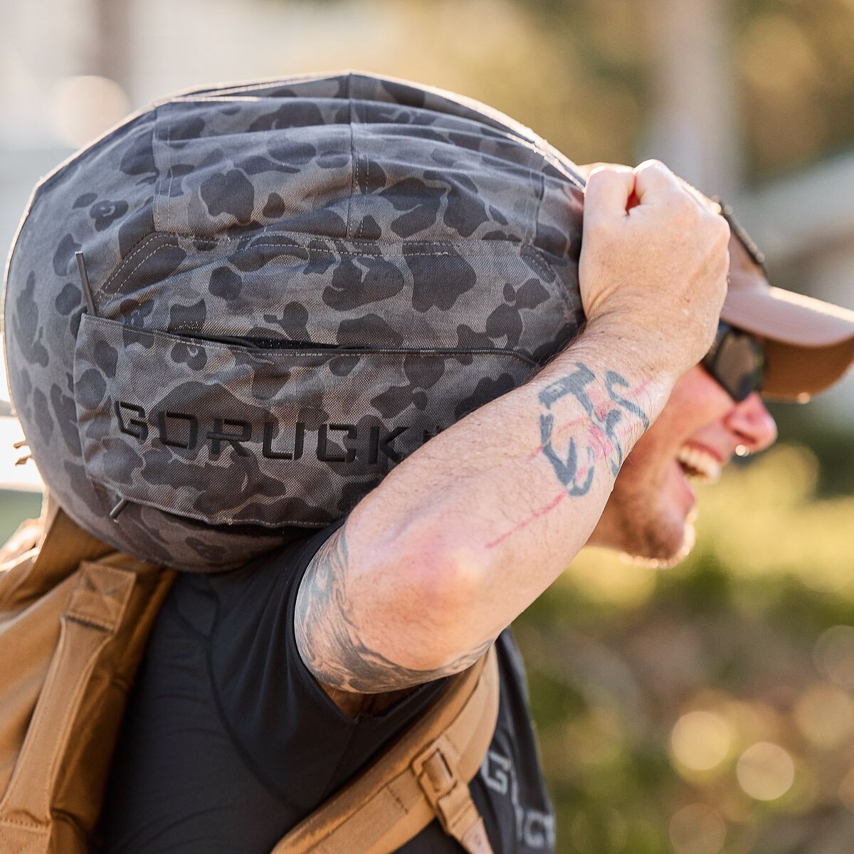 Man outdoors lifting a camo GORUCK fitness sandbag, wearing sunglasses and a brown cap