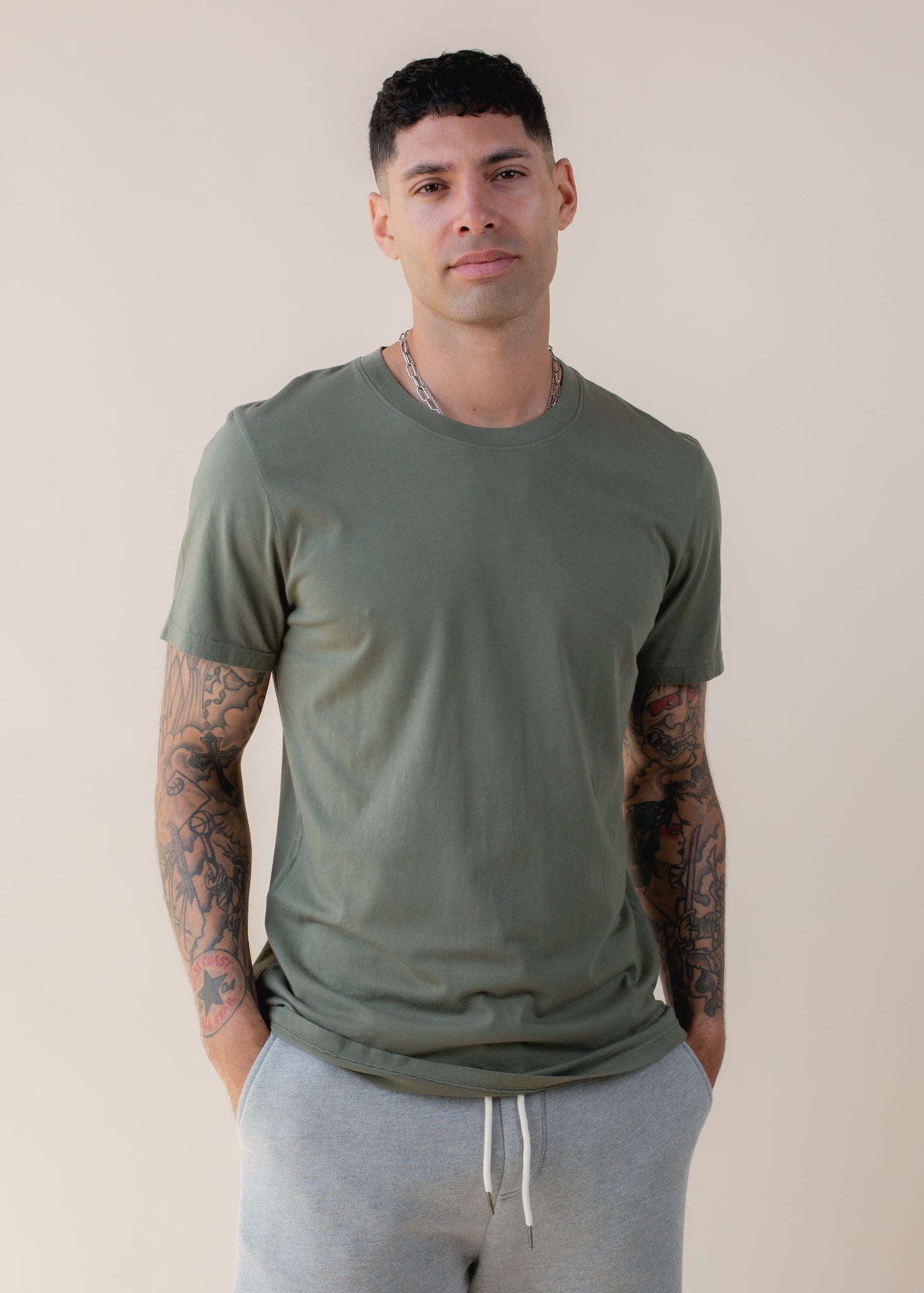 Man wearing an artichoke green basic tee and grey joggers, standing against a neutral background