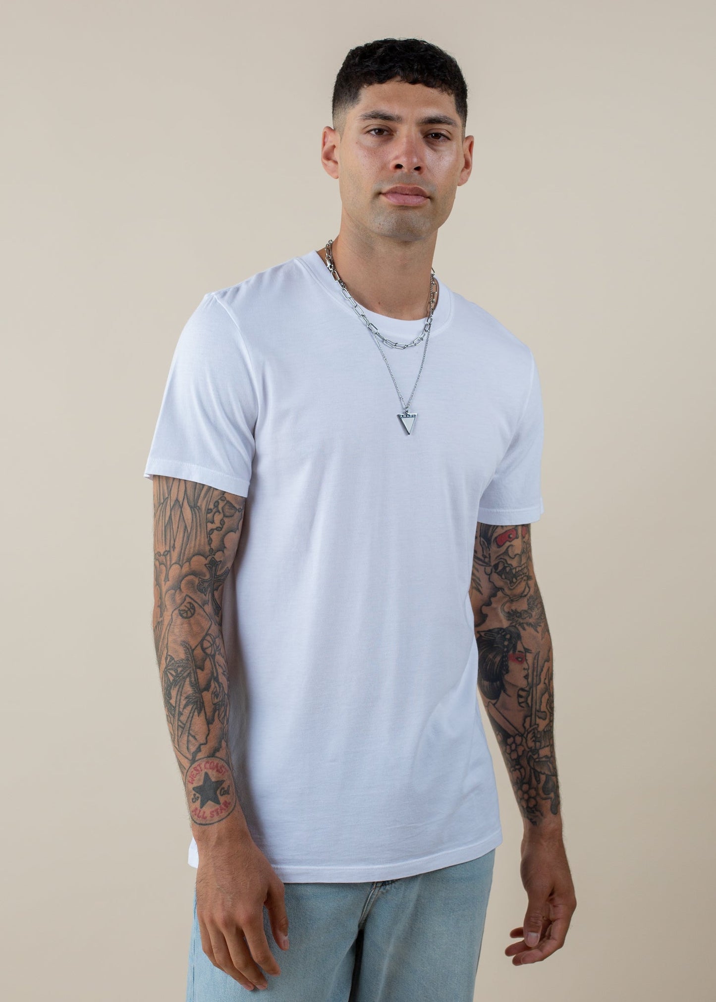 Man wearing basic white tee, light jeans, silver necklaces, and showing tattooed arms