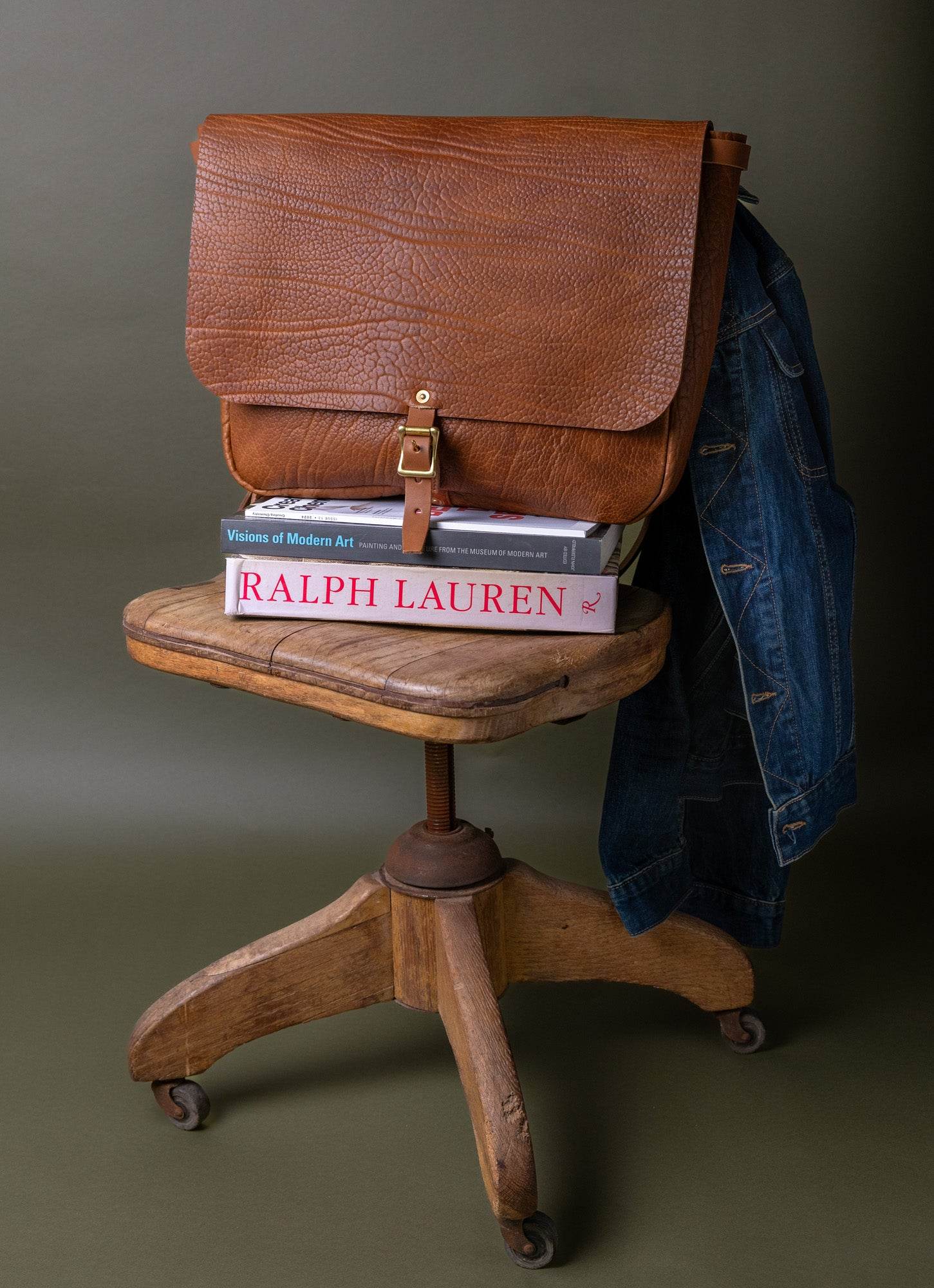 Brown leather messenger bag on stacked books and denim jacket draped over wooden chair, men's lifestyle
