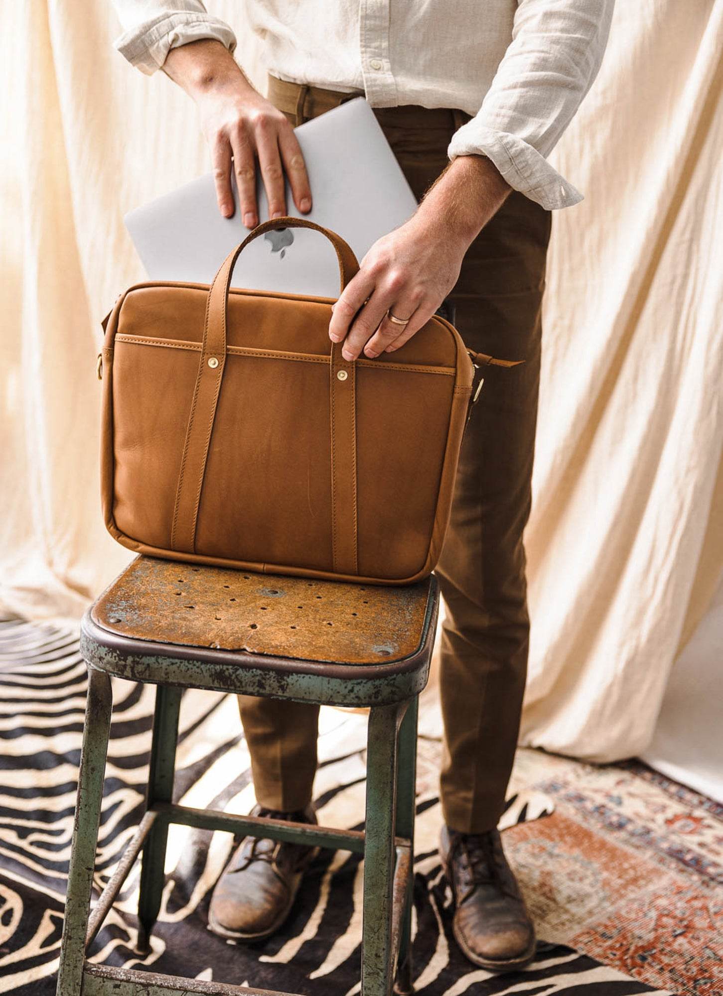 Man with laptop placing it in brown leather briefcase, stylish men's fashion on rustic stool