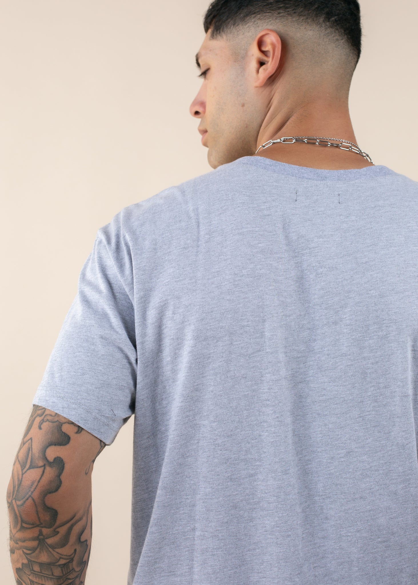 Man wearing oversized heather grey tee, chain necklace, and showing arm tattoo