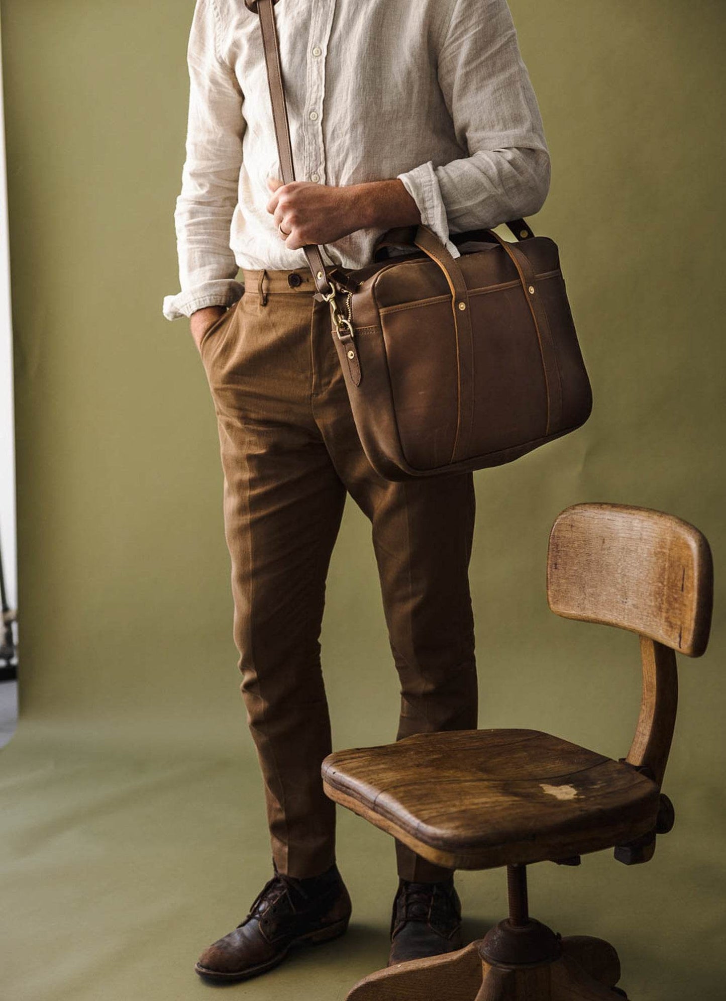 Stylish man in brown pants and white shirt with leather messenger bag, vintage wood chair, lifestyle fashion