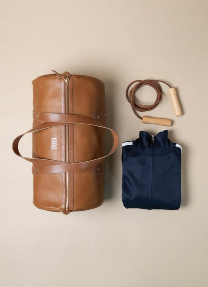 Men's gym essentials with brown leather duffel bag, navy zip-up jacket, and jump rope.