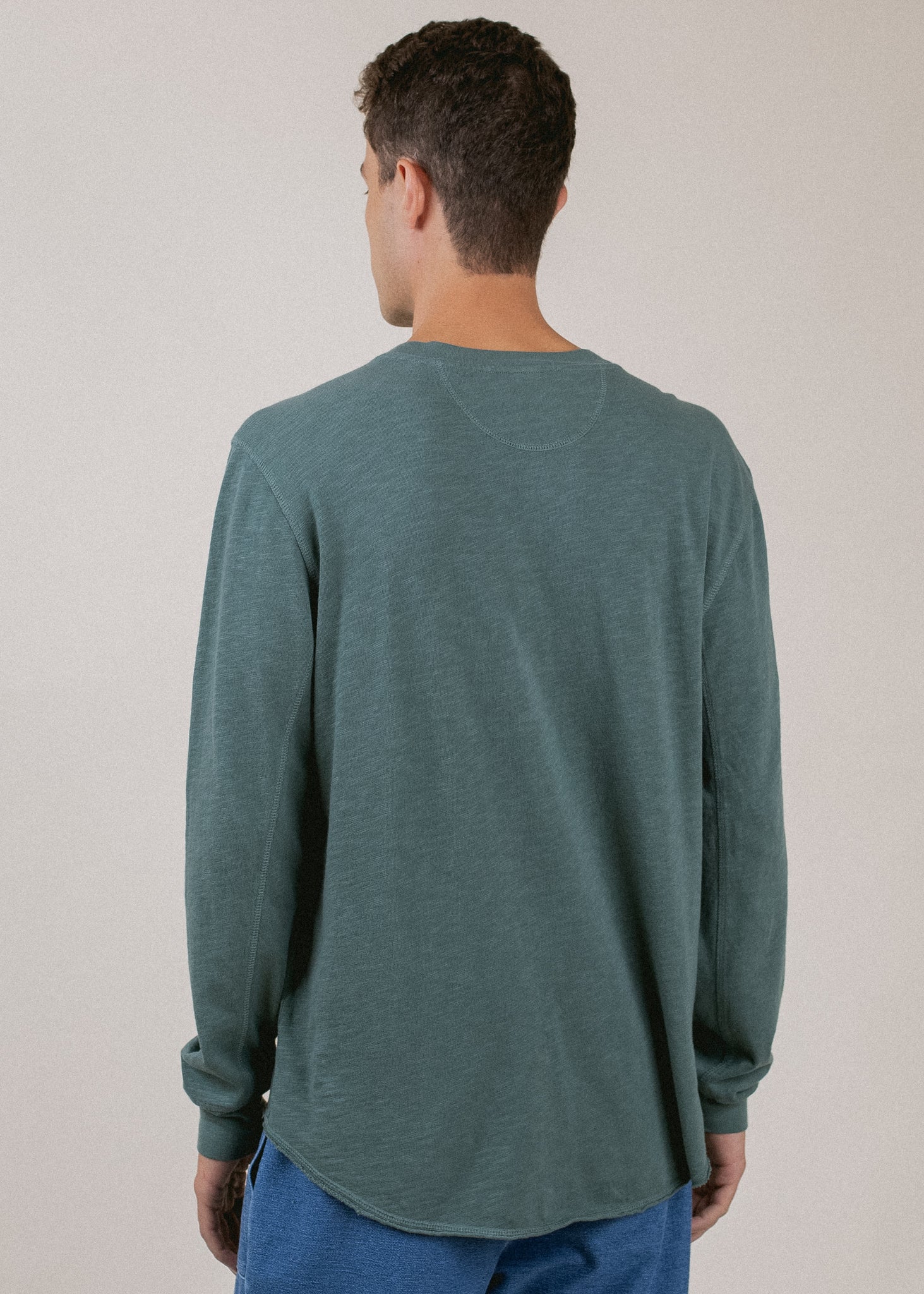 Man wearing a pine green Henley long sleeve shirt, viewed from the back, on neutral background