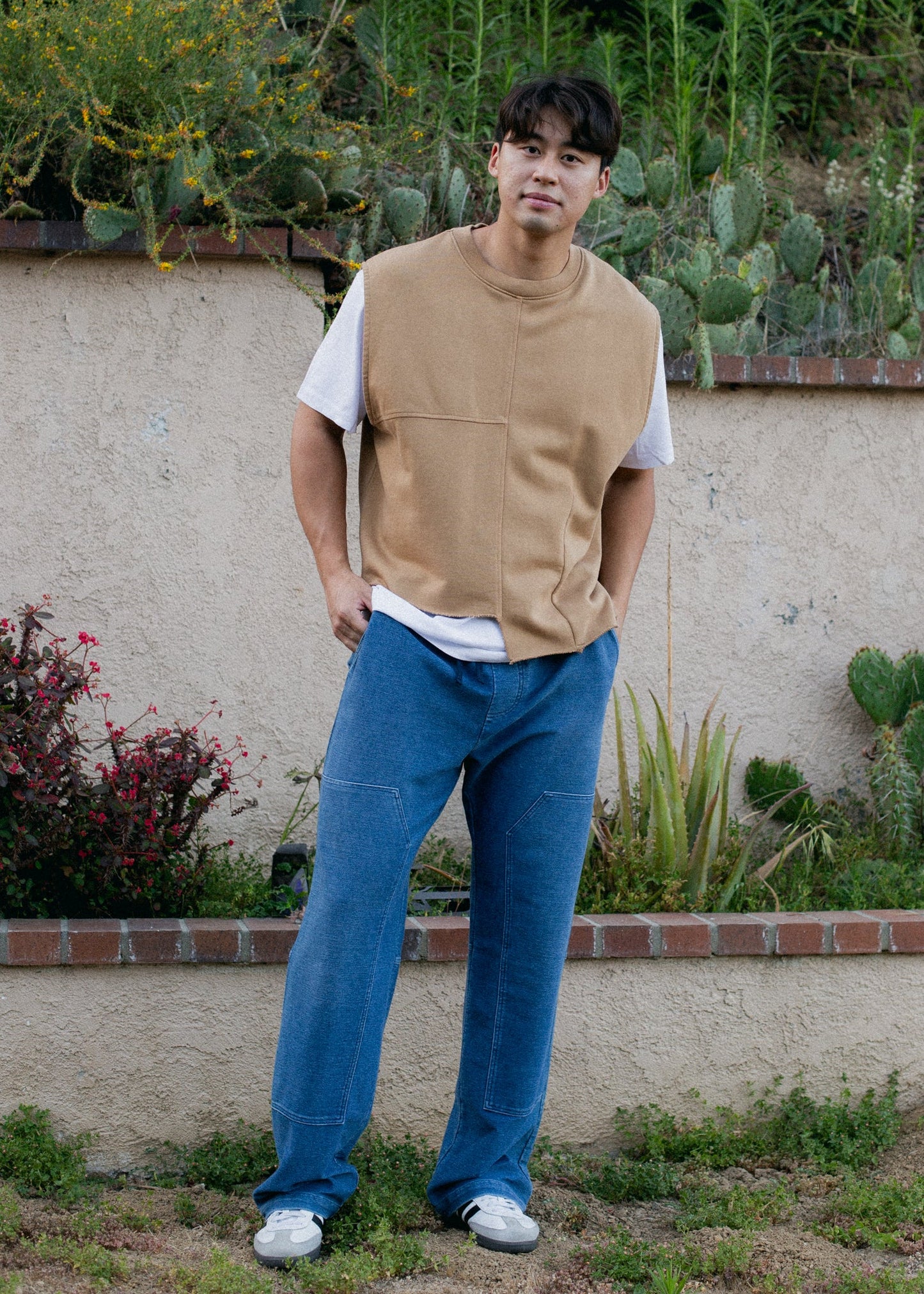 Man wearing indigo patch denim trousers and beige vest standing outdoors by a garden wall