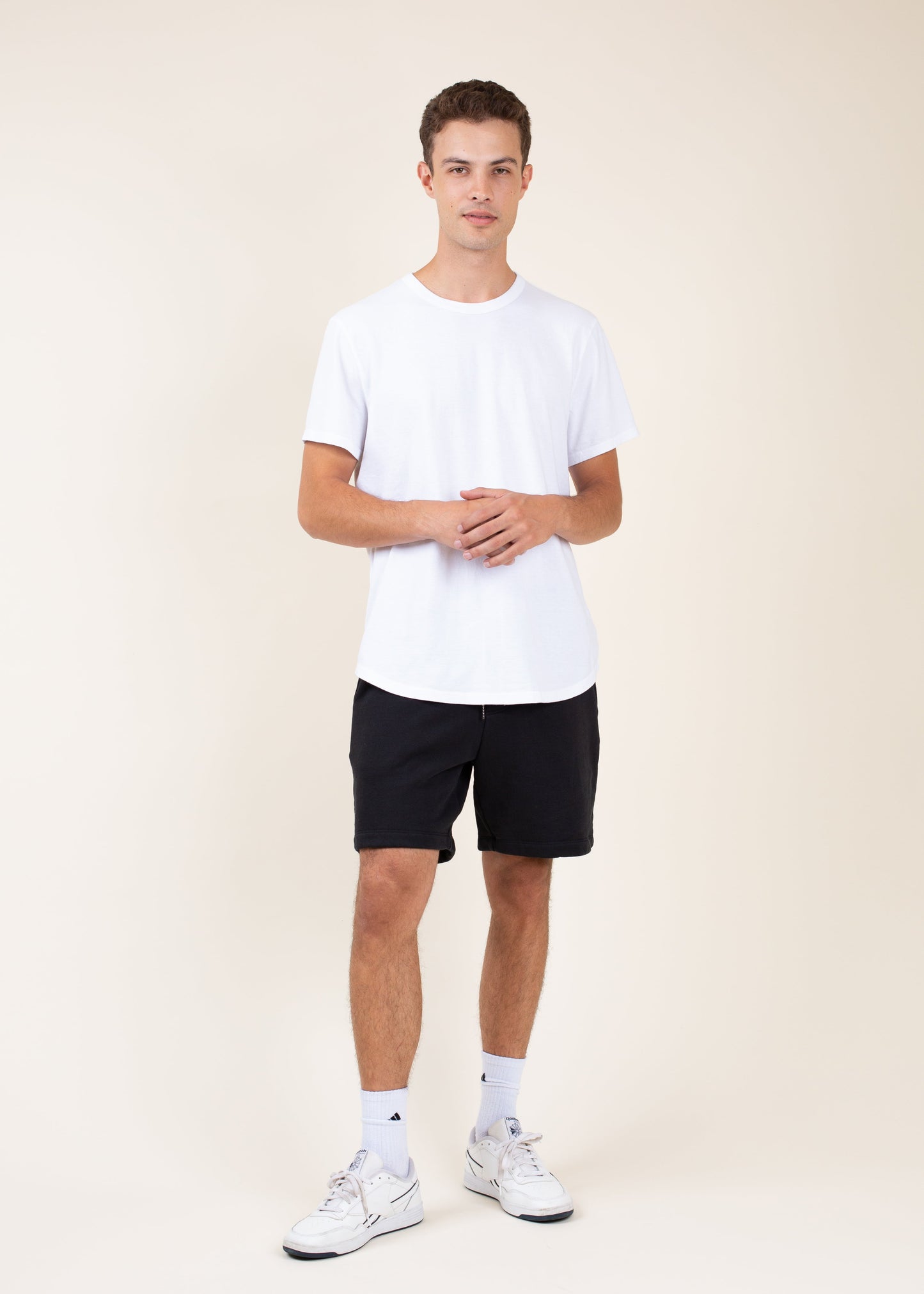 Man in white t-shirt, black shorts, and sneakers posing against neutral background for MANN fashion