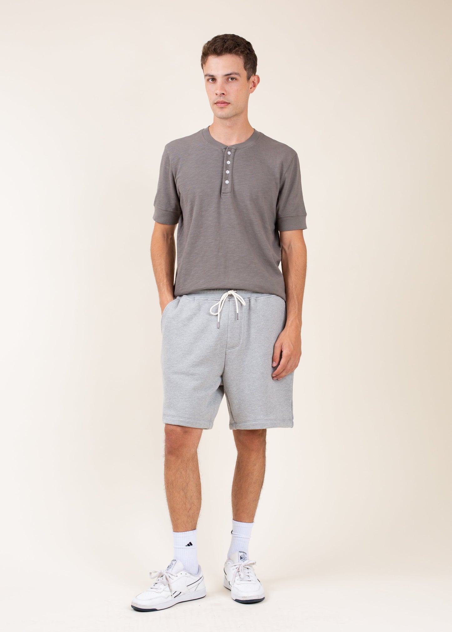 Man in casual grey henley shirt and light grey drawstring shorts, white sneakers, studio background