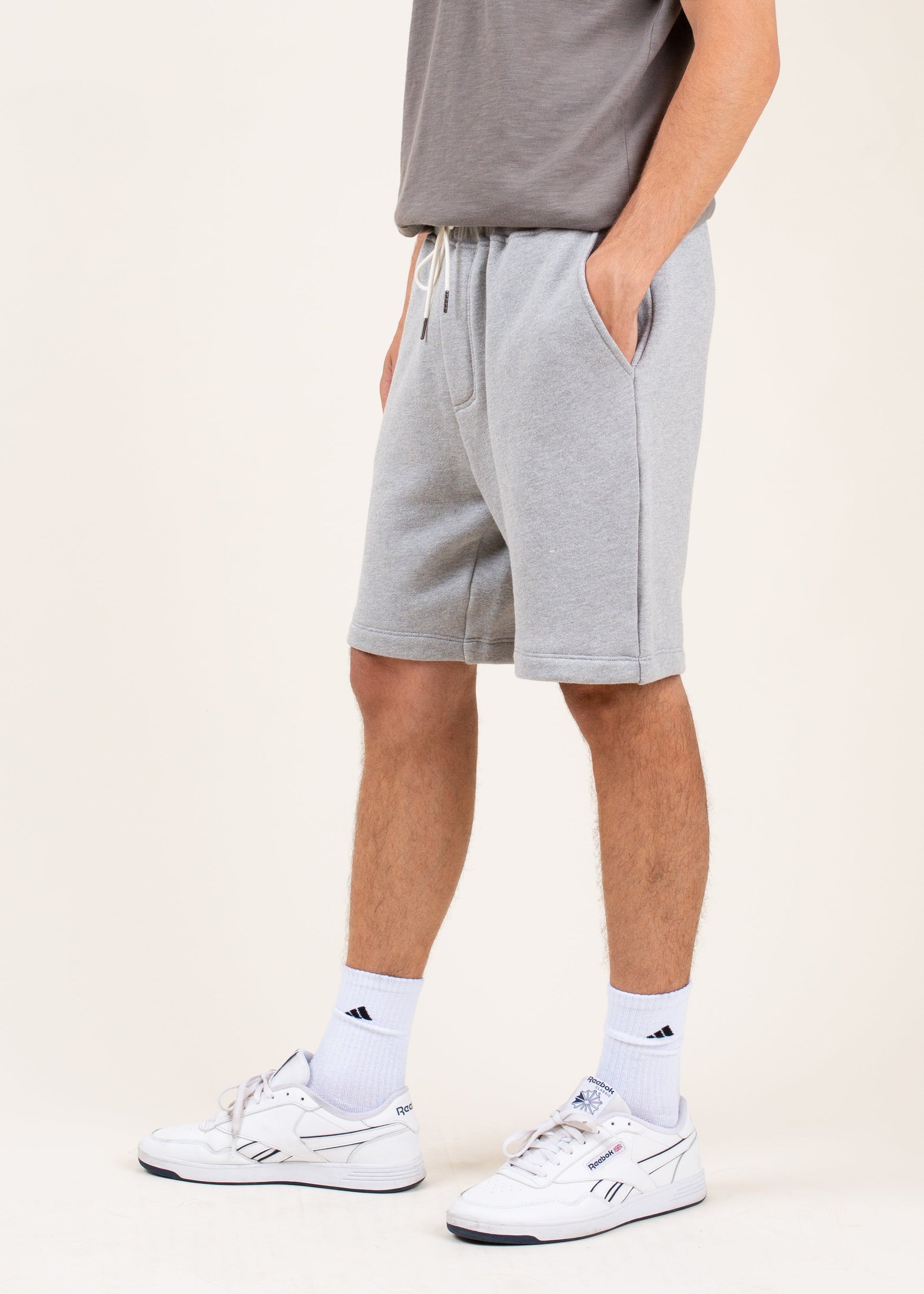 Man wearing grey athletic shorts, white Reebok sneakers, and white crew socks on neutral background