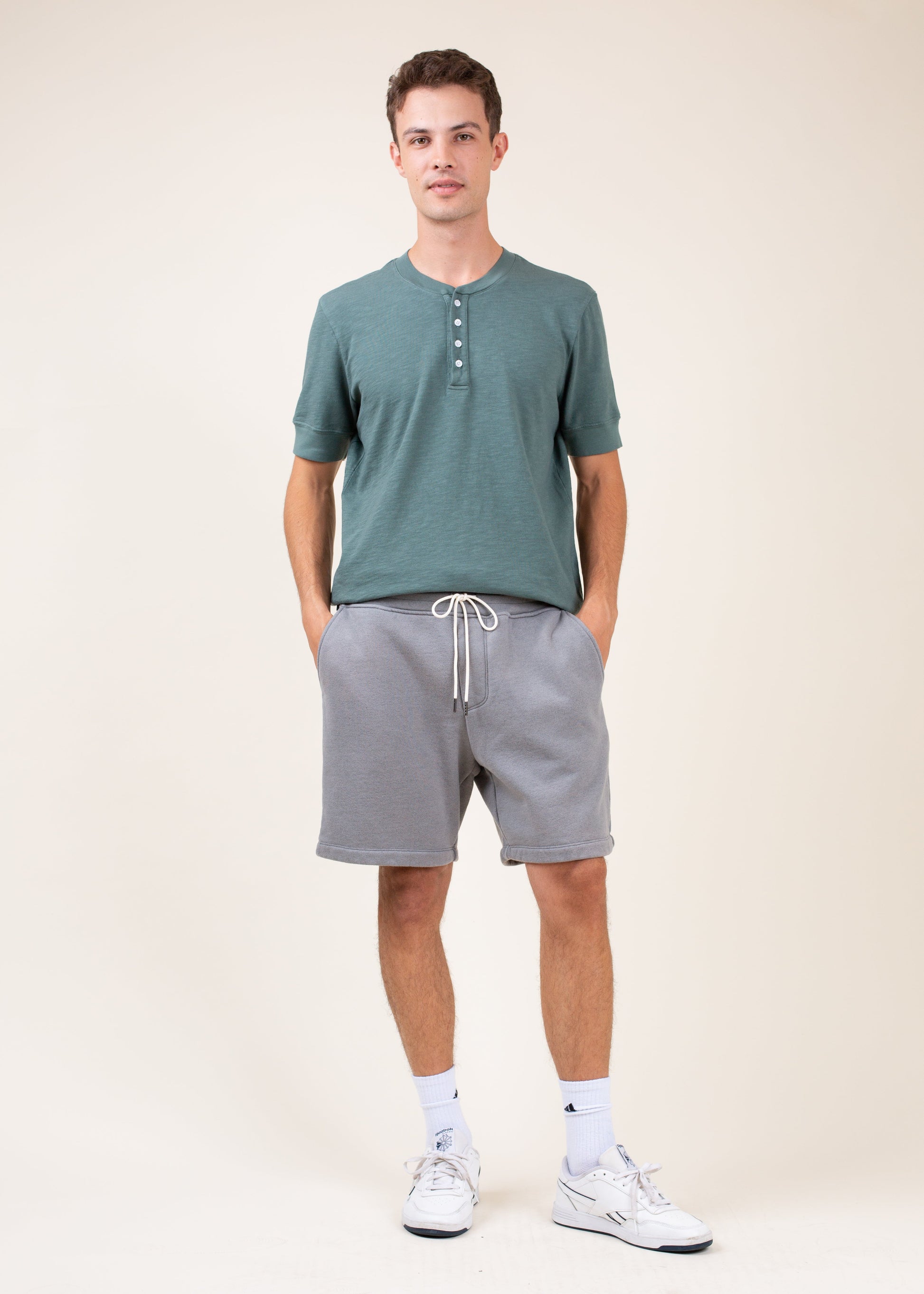 Man wearing green henley shirt, gray drawstring shorts, and white sneakers, casual menswear style