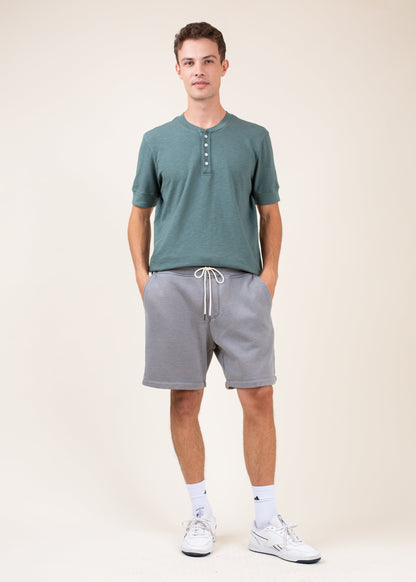 Man wearing green henley shirt, gray drawstring shorts, and white sneakers, casual menswear style