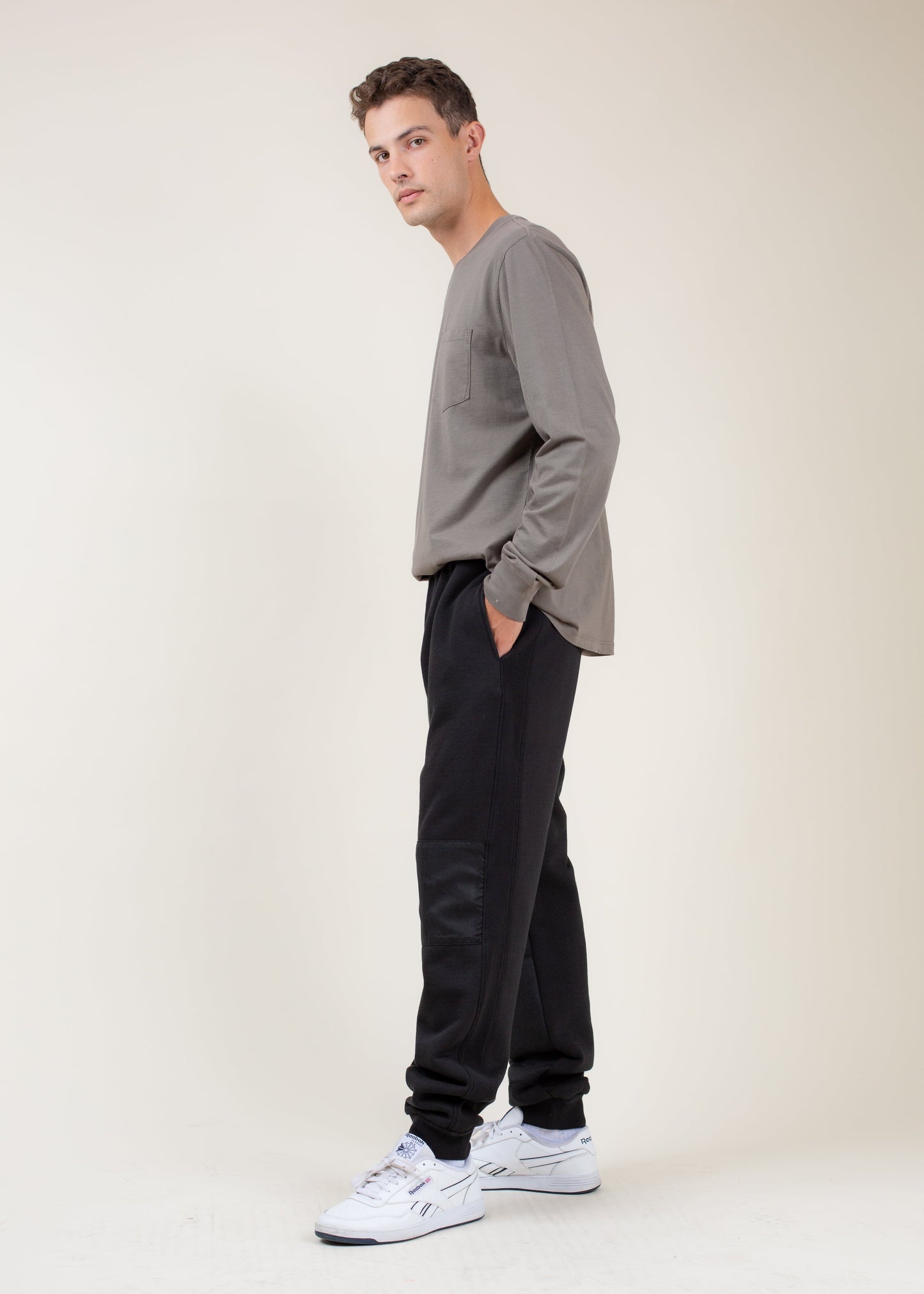 Man in casual grey long sleeve shirt and black joggers with white sneakers on neutral background