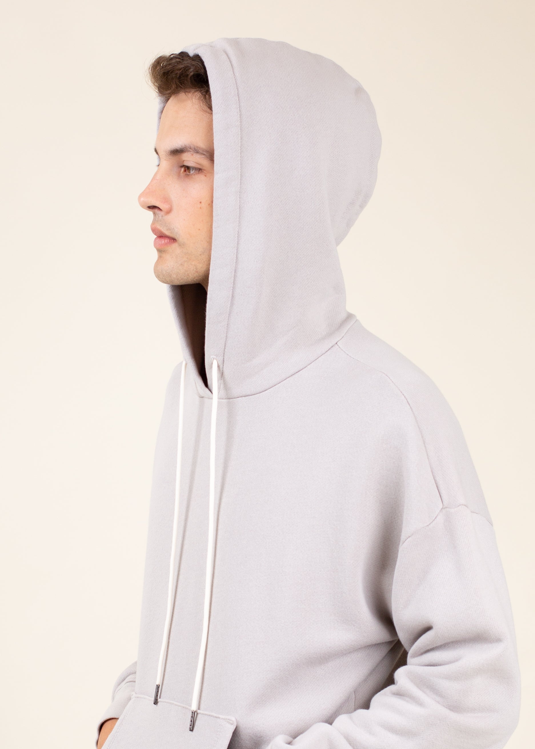 Man in light gray hoodie with white drawstrings, side view, minimalist background, MANN fashion.