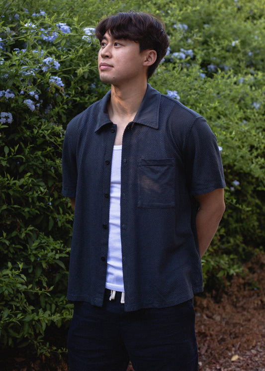 Man in navy mesh button-down shirt and white tee standing outdoors by green bushes