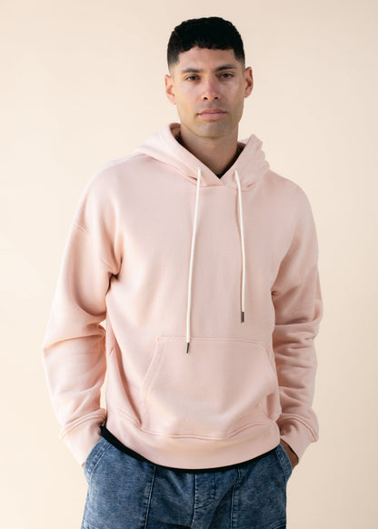 Man in light pink hoodie and denim pants posing against beige background for MANN fashion