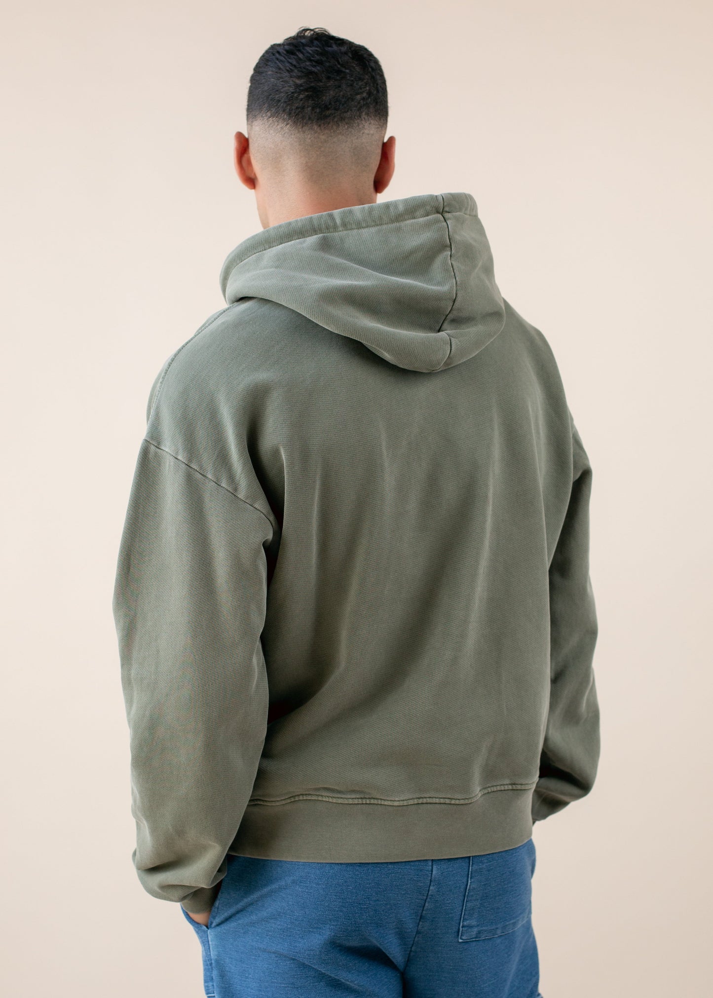 Man wearing olive green hoodie and blue pants, back view, neutral background, men's fashion