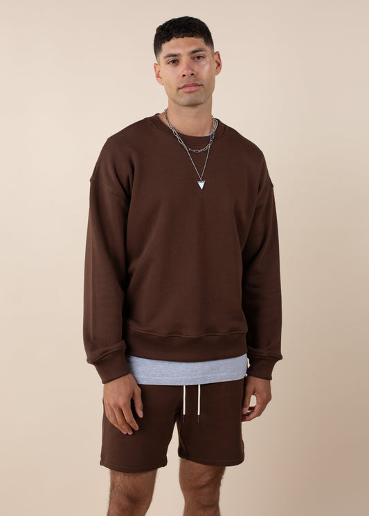Man in brown sweatshirt and shorts, layered with gray shirt, wearing silver necklace, neutral backdrop