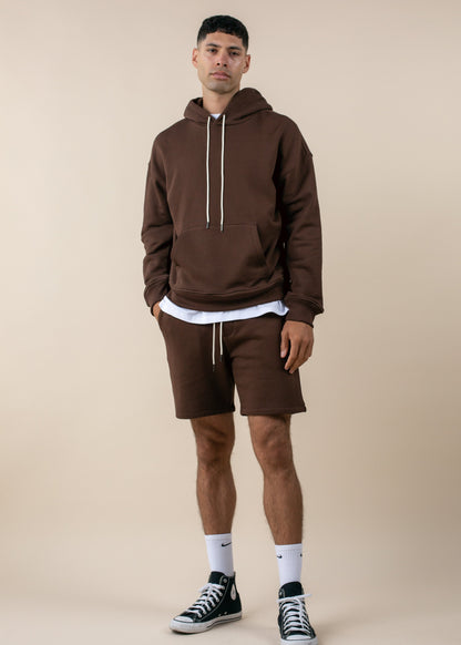 Man in brown hoodie and shorts, white socks, and black sneakers, posing against beige background for MANN men's fashion marketplace.