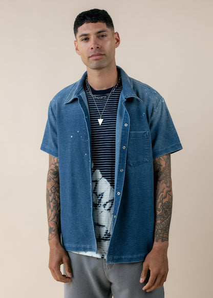 Man in casual blue short-sleeve shirt, striped tee, silver necklace, tattoos, neutral background