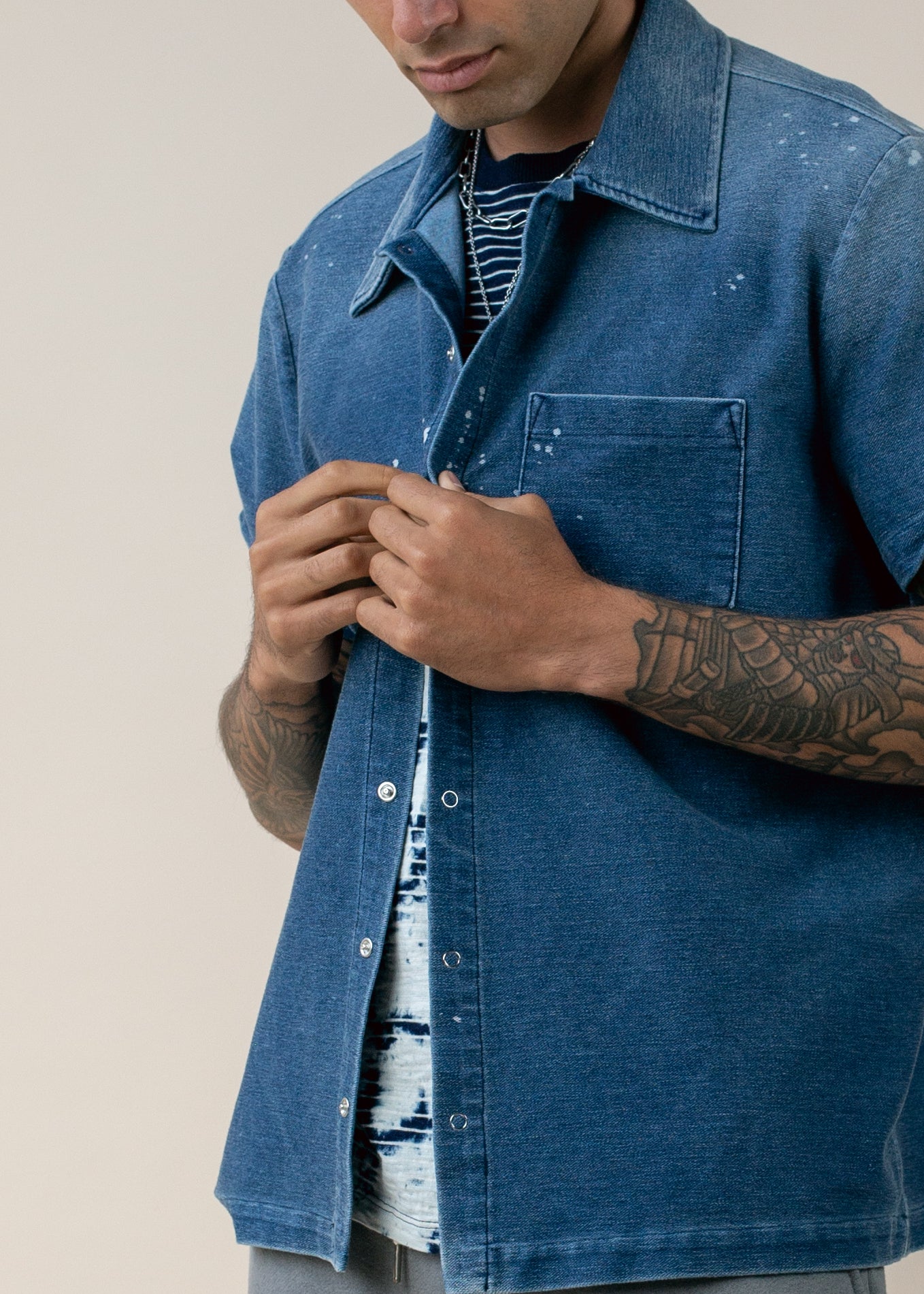 Man wearing a blue short sleeve denim shirt, striped tee, silver chain, tattooed arm