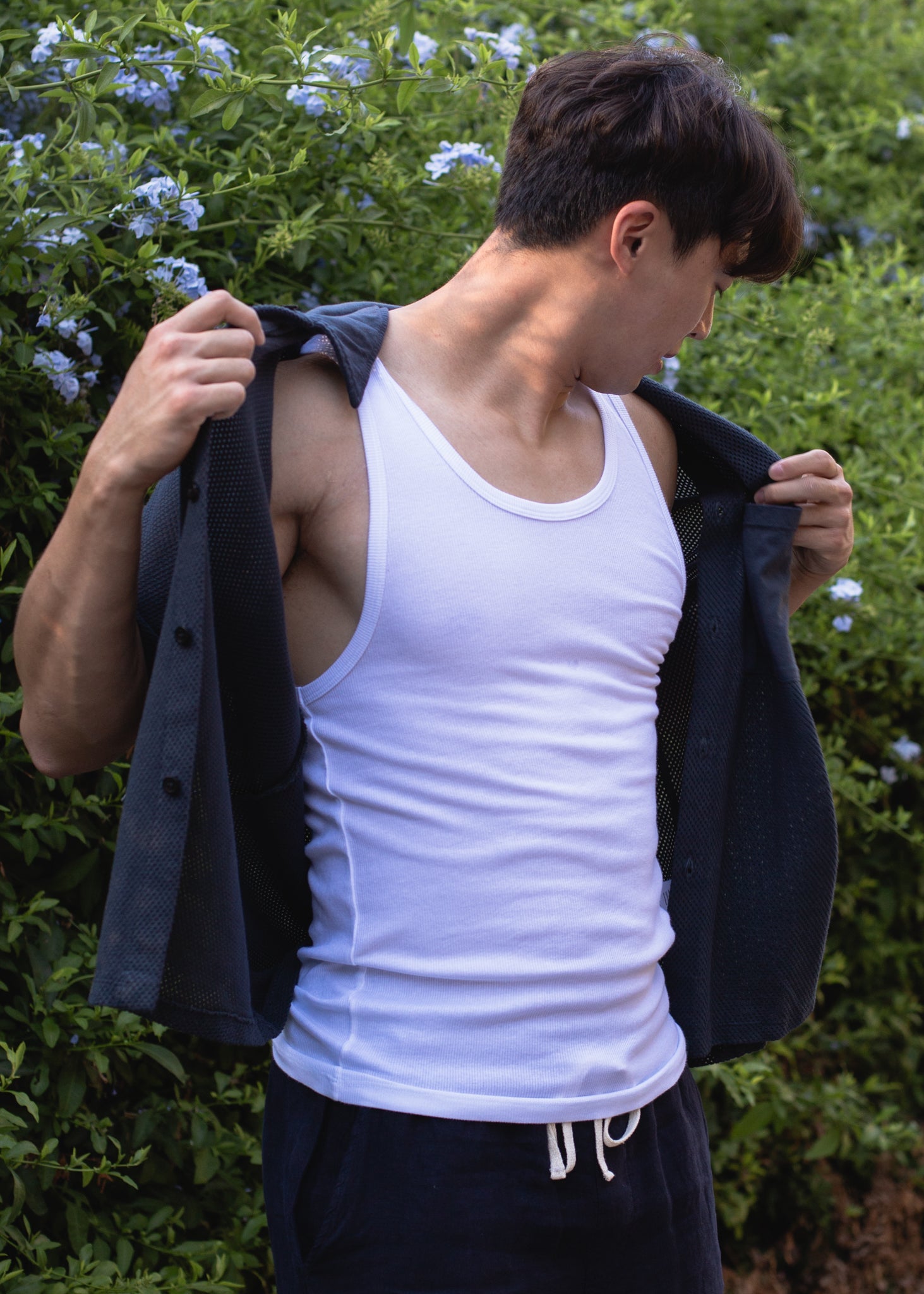 Man in white ribbed tank top and dark open shirt standing outdoors with greenery and flowers